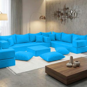Może przedstawiać: Duża, modułowa, turkusowa sofa narożna z wieloma poduszkami i pufami. Sofa jest ustawiona w kształcie litery L, o niskim profilu i nowoczesnym designie. Drewniany stolik kawowy i dekoracyjne wazony znajdują się przed sofą.