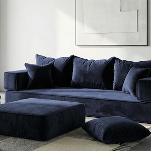 May include: A contemporary navy blue velvet sofa with a matching ottoman and cushions. The sofa has a low profile and a minimalist design. The square ottoman and cushions complement the sofa. The furniture is placed on a patterned rug. The sofa is approximately 200 cm in length.