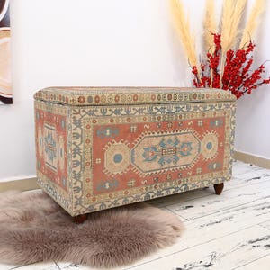 May include: A rectangular storage ottoman with a colorful patterned fabric top. The fabric features a traditional rug design with red, blue, and beige colors. The ottoman is sitting on a light brown rug.