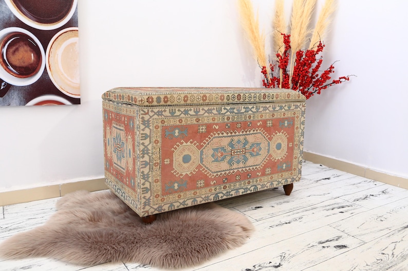 May include: A rectangular storage ottoman with a colorful patterned fabric top. The fabric features a traditional rug design with red, blue, and beige colors. The ottoman is sitting on a light brown rug.