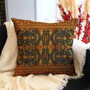 May include: A decorative throw pillow with a dark green and gold geometric pattern, accented with red and orange details. The pillow is rectangular and rests on a white blanket, suggesting a home decor theme.