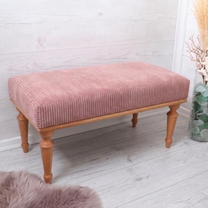 May include: A pink corduroy bench with a light wood frame and legs. The bench is rectangular and has a soft, textured appearance. The legs are turned and add a touch of elegance. The bench is placed on a light wood floor.