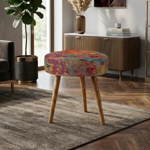 Può includere: Uno sgabello rotondo caratterizzato da un vivace motivo astratto multicolore nei toni dell'arancione, del blu e del rosa sulla seduta. Lo sgabello è sostenuto da tre gambe in legno chiaro inclinate verso l'esterno per una base stabile.