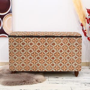Può includere: Un pouf vintage con funzione di contenitore, con un motivo geometrico in tonalità di marrone, arancione e bianco. Il pouf ha una struttura e gambe in legno.