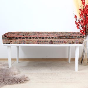 May include: White bench with a patterned rug cushion. The bench has four legs and is made of wood. The rug cushion is a traditional design with red, blue, and white colors.