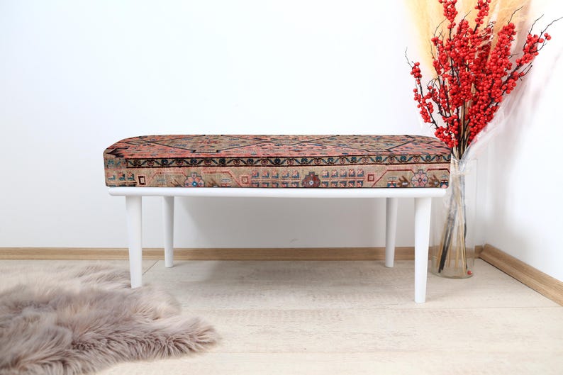 May include: White bench with a patterned rug cushion. The bench has four legs and is made of wood. The rug cushion is a traditional design with red, blue, and white colors.