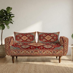 Może przedstawiać: Dwuosobowa sofa z tapicerką z tkaniny w czerwone, niebieskie i beżowe wzory przypominające tradycyjny dywan. Sofa posiada dwie prostokątne poduszki oparciowe, płaskie siedzisko oraz zaokrąglone drewniane nóżki w kolorze ciemnego brązu.