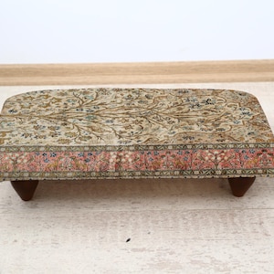 May include: A rectangular footstool with a patterned fabric top. The fabric features a beige background with floral and vine designs in shades of brown, blue, and green. The bottom edge has a pink and blue floral border. The stool is 24 inches long.
