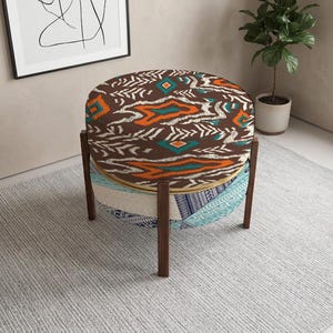 May include: A round ottoman featuring a brown top with an orange and teal geometric pattern. The base is supported by four dark wooden legs and includes a lower shelf upholstered in a light blue and white patterned fabric with dark blue accents.
