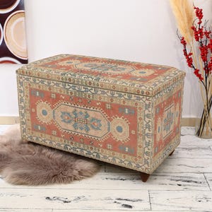 May include: A decorative storage ottoman with a colorful patterned fabric top. The fabric features a traditional rug design with red, blue, and cream colors.