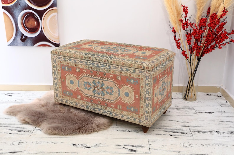 May include: A decorative storage ottoman with a colorful patterned fabric top. The fabric features a traditional rug design with red, blue, and cream colors.