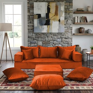 May include: An orange velvet sofa with matching throw pillows and an ottoman. The living room features a stone fireplace, a patterned rug, and a large abstract painting. A floor lamp and a potted plant add to the decor.
