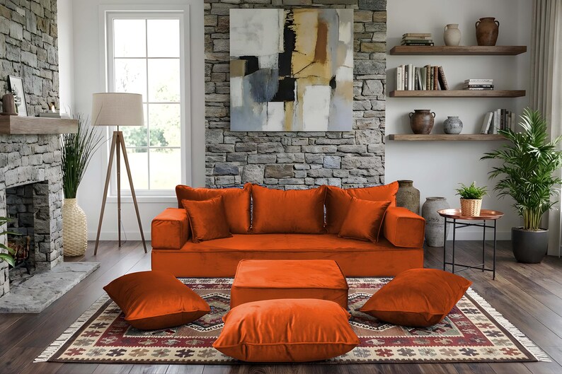 May include: An orange velvet sofa with matching throw pillows and an ottoman. The living room features a stone fireplace, a patterned rug, and a large abstract painting. A floor lamp and a potted plant add to the decor.