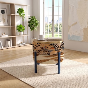 May include: A round ottoman with a light wood top and a patterned fabric base featuring black, tan, and brown animal print designs. The ottoman is supported by four dark blue metal legs and is topped with a white ceramic mug and a book.