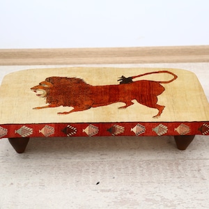 Puede incluir: Un taburete rectangular de madera con una parte superior beige que presenta una ilustración detallada de un león en tonos marrones y rojos. El taburete tiene un borde rojo con patrones decorativos y patas marrones oscuras.
