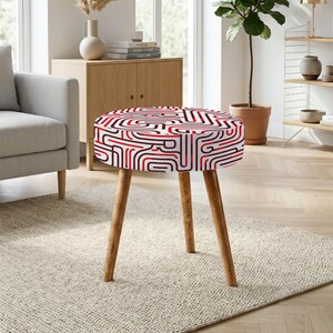 Könnte beinhalten: Ein runder Hocker mit drei hellbraunen Holzbeinen und einer gepolsterten Sitzfläche, die mit einem geometrischen Muster aus ineinandergreifenden roten, dunkelroten und weißen Linien versehen ist, welche ein labyrinthartiges Design bilden.