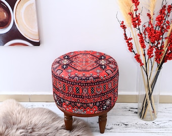 May include: A round red and black patterned ottoman with wooden legs. The ottoman is sitting on a white floor with a fluffy rug in the foreground.