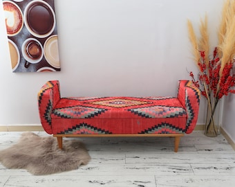 Könnte beinhalten: Eine rote Bank mit einem geometrischen Muster in Blau-, Grün- und Gelbtönen. Die Bank hat Holzbeine und Armlehnen. Ein flauschiger, grauer Teppich liegt vor der Bank. Eine Leinwand mit Kaffee-Kunst hängt an der Wand.