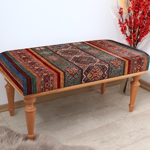 May include: A wooden bench with a colorful patterned cushion. The cushion has a geometric design with shades of red, blue, green, and yellow. The bench has four wooden legs.