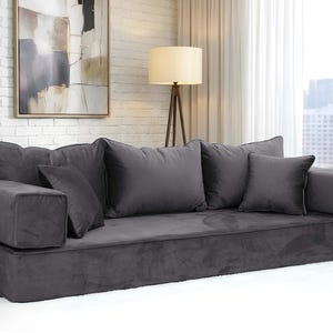 May include: A dark gray, low-profile sofa with multiple plush cushions. The sofa has a rectangular base and armrests, and is placed in front of a window with a cityscape view. The room has a neutral color palette.