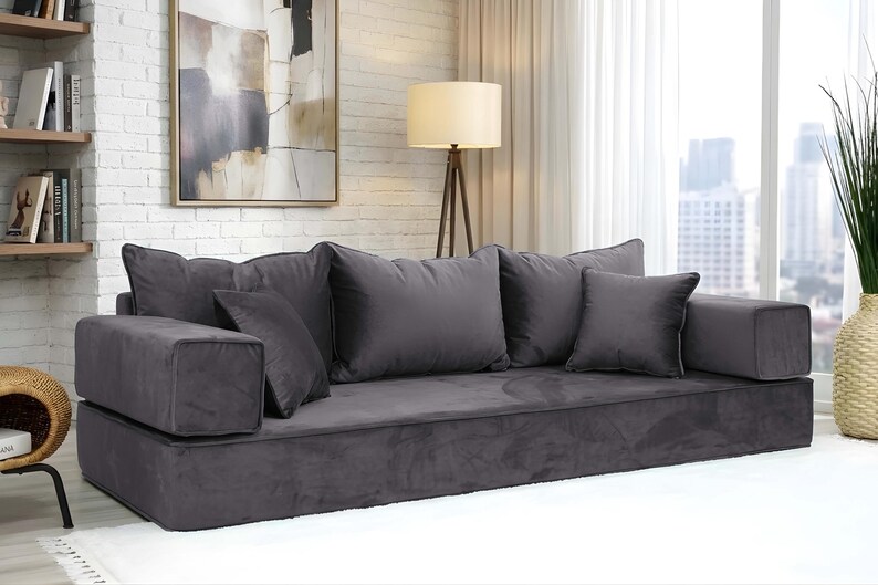 May include: A dark gray, low-profile sofa with multiple plush cushions. The sofa has a rectangular base and armrests, and is placed in front of a window with a cityscape view. The room has a neutral color palette.