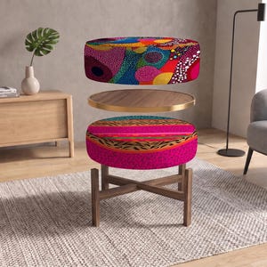 Könnte beinhalten: Ein runder Hocker mit Holzgestell und gekreuzten Beinen, der ein lebhaftes, mehrfarbig gepunktetes Muster auf dem oberen Kissen und ein leuchtend rosa, orangefarbenes und tierisches Druckdesign auf dem unteren Kissen aufweist, getrennt durch ein Holztablett.