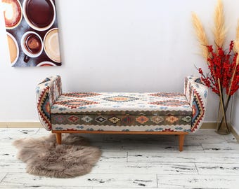 May include: A bench with a patterned fabric seat and curved armrests. The fabric features a geometric design in shades of red, blue, and beige. The bench has wooden legs and sits on a light-colored wooden floor. A fluffy rug is in front of the bench.