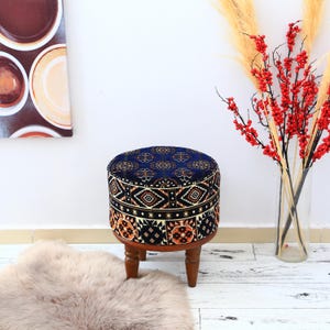 Può includere: Un pouf rotondo imbottito con un top e lati a fantasia blu scuro. Il motivo include forme geometriche arancioni, bianche e nere. Il pouf ha una base in legno con quattro gambe corte e tornite. Un soffice tappeto marrone chiaro è in primo piano.