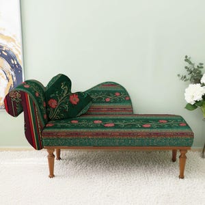 May include: A decorative chaise lounge with a floral pattern in green, red, and gold. The chaise has a curved back and armrest, with wooden legs. The background includes a painting and a vase of white flowers.