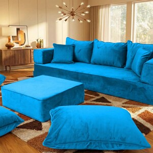 May include: A modern living room setup featuring a large, bright blue modular sofa with matching throw pillows and a square ottoman. Several large floor cushions and potted plants add to the room's inviting atmosphere. The room has a warm, inviting feel.