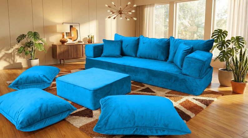 May include: A modern living room setup featuring a large, bright blue modular sofa with matching throw pillows and a square ottoman. Several large floor cushions and potted plants add to the room's inviting atmosphere. The room has a warm, inviting feel.
