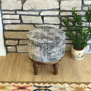 May include: A round, upholstered ottoman with a dark gray and white pattern, set on four wooden legs. The ottoman is placed on a wooden floor, near a small potted plant and a stone brick wall. The ottoman is a decorative furniture piece.