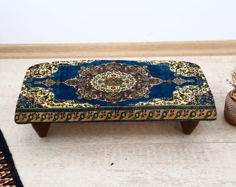 May include: A rectangular footstool with a dark blue, patterned top featuring a central medallion design in brown, gold, and cream. The stool has short, dark wooden legs and is placed on a light-colored floor.