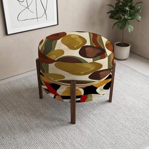 May include: A round ottoman featuring a multi-colored abstract pattern with organic shapes in shades of brown, olive green, mustard yellow, and cream. The seat is supported by four slender, dark brown wooden legs arranged in a vertical frame.