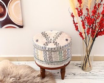 May include: A round, white and beige patterned ottoman with wooden legs. The ottoman is sitting on a white rug with a fluffy texture.