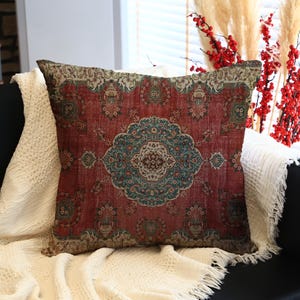 May include: A cushion with a vintage rug design. The cushion has a red background with a central medallion in teal and brown. It is placed on a black sofa and a cream-coloured blanket. The cushion measures approximately 40cm x 40cm.