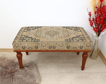 May include: A wooden ottoman with a beige and blue patterned fabric top. The ottoman has four turned legs.