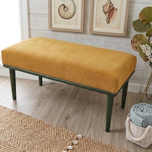 May include: A mustard yellow upholstered bench with a corduroy texture. The bench has a dark green frame and legs. Decorative items include framed seashell art, a woven basket with a rolled blanket, and a natural fiber rug.
