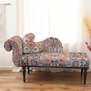 May include: A chaise lounge with a patterned fabric in shades of orange, blue, and cream. The chaise has a curved back and armrest, with black legs. A decorative pillow matches the fabric. The room also features a macrame wall hanging and a small side table.