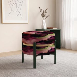 May include: A round ottoman featuring a vibrant abstract pattern in shades of deep purple, magenta, and soft green. The seat is accented by a thin gold-toned metal band and is supported by four dark green legs that extend upward to the seat frame.