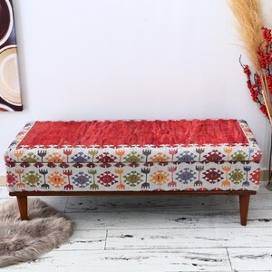 May include: A rectangular ottoman with a red, textured top and a patterned fabric base. The base features a repeating geometric design in various colors. The ottoman has wooden legs and is placed on a white wooden floor.