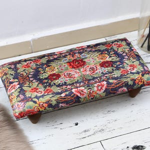 Könnte beinhalten: Ein Fußhocker mit floralem Muster und Holzbeinen. Der Fußhocker hat einen dunkelblauen Hintergrund mit roten, rosa und gelben Blumen.