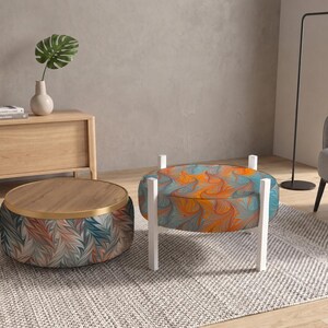 May include: A round ottoman with a blue and orange marbled pattern and white legs, positioned next to a low round table featuring a wooden top, a gold rim, and a base upholstered in a blue, orange, and grey leaf-like patterned fabric.