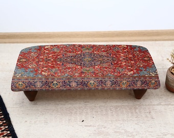 May include: A rectangular, low wooden table with a patterned rug top. The rug features a red, blue, and cream floral design. The table has four short, dark brown legs and is suitable for a living room or bedroom.