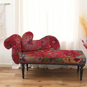Puede incluir: Una chaise longue roja con un estampado floral, con un cojín en forma de corazón y un reposabrazos enrollado. El sofá tiene patas negras y está sobre una cortina blanca y un suelo de madera. Elementos decorativos: macramé de pared y mesita auxiliar.