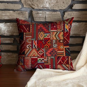 Puede incluir: Cojín decorativo cuadrado con un diseño de patchwork en tonos rojos, naranjas y marrones. El cojín presenta patrones y formas geométricas. El cojín está colocado sobre una superficie de madera frente a una pared de piedra y una manta color crema.