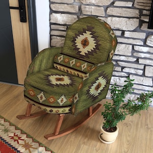 May include: A green patterned rocking chair with a matching lumbar pillow. The chair has a wooden frame and is placed on a wooden floor. A small potted plant sits nearby. The wall behind the chair is made of stone.