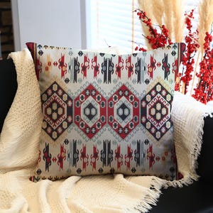 May include: A decorative throw pillow with a geometric pattern in red, black, and gray on a cream background. The pillow is placed on a black sofa with a cream-colored blanket.