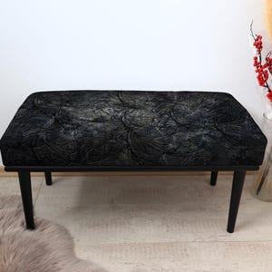 May include: A black bench with a rectangular seat and four black legs. The seat has a textured, patterned design in shades of black and gray. The bench is a piece of furniture.
