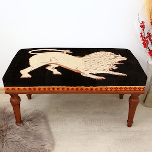 Pode incluir: Um pufe retangular com um tampo preto com um desenho de leão bege. O pufe tem uma borda decorativa e assenta em quatro pernas de madeira. O design geral é vintage e decorativo.